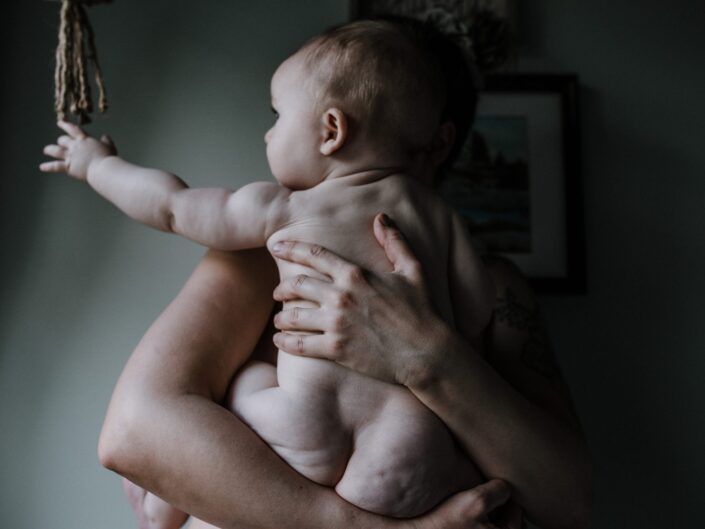 A naked baby is held by his mother, as he reaches for something out of the frame.